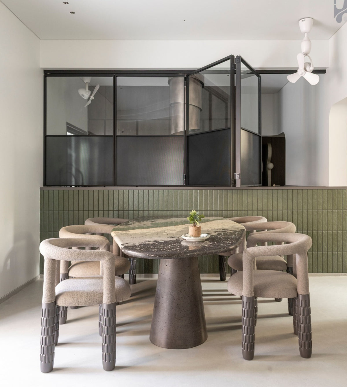 A modern dining room setup featuring a round dark stone-top table with a sculptural pedestal base, surrounded by six upholstered dining chairs with curved backs and textured gray lower legs. The room has a light green ribbed tile wall behind the table, black-framed glass partition panels, and a white ceiling with a decorative ceiling fan.