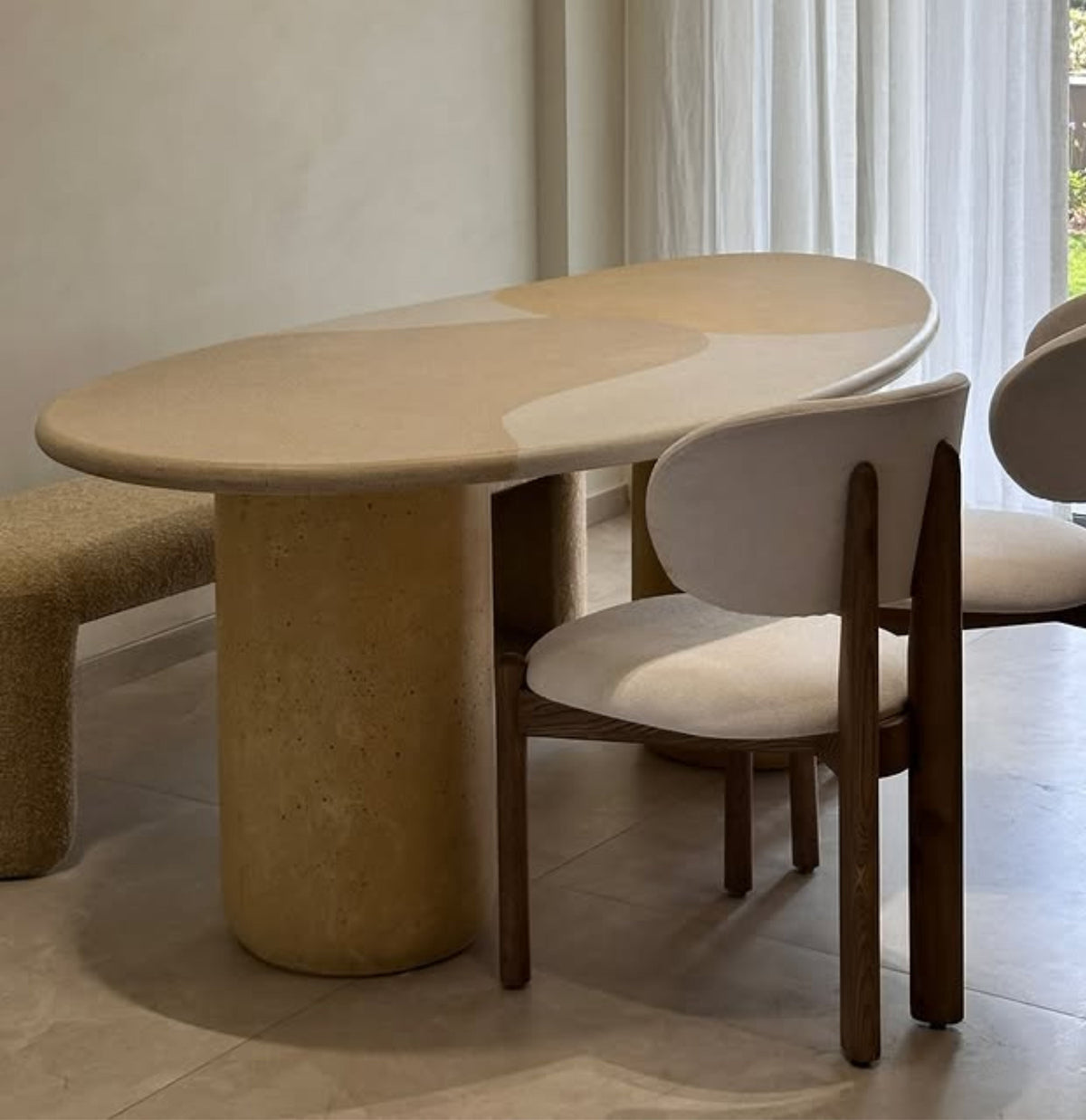 An oval dining table with a textured beige stone top and cylindrical base, paired with upholstered wooden chairs in a bright, modern interior.